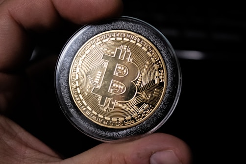 A close-up view of a hand holding a gold-colored coin with the Bitcoin logo prominently displayed in the center. The coin is encased in a protective transparent cover, and intricate circuit-like patterns are etched around the logo, symbolizing digital technology.