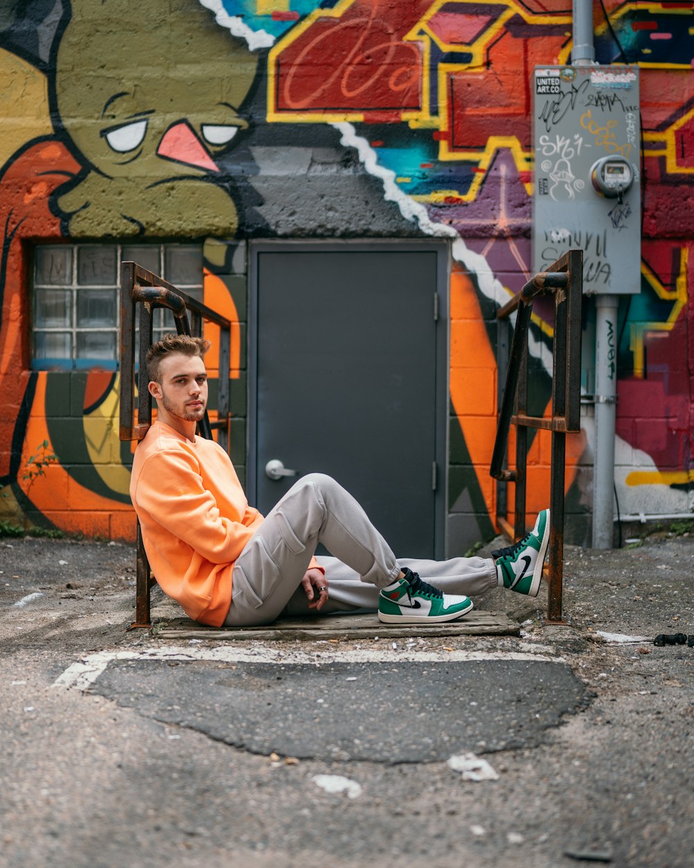 uomo in camicia arancione a maniche lunghe e pantaloni grigi seduto sul pavimento di cemento grigio
