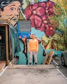 A man stands on a wooden platform in front of a vibrant mural featuring a large face and red flowers. The wall is covered with colorful graffiti including greens, blues, and reds. Nearby are some plants and a parked car, creating a contrast between urban art and natural elements.