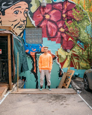 A man stands on a wooden platform in front of a vibrant mural featuring a large face and red flowers. The wall is covered with colorful graffiti including greens, blues, and reds. Nearby are some plants and a parked car, creating a contrast between urban art and natural elements.