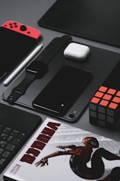 A neatly arranged assortment of tech gadgets and accessories displayed on a gray surface. The layout includes a red and black handheld gaming console, a smartwatch with a black strap, a black smartphone, a white wireless earbud case, a black stylus, and a Rubik's cube. A part of a comic book featuring a superhero is partially visible at the bottom.