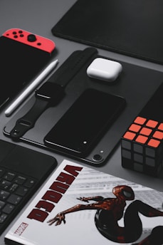 A neatly arranged assortment of tech gadgets and accessories displayed on a gray surface. The layout includes a red and black handheld gaming console, a smartwatch with a black strap, a black smartphone, a white wireless earbud case, a black stylus, and a Rubik's cube. A part of a comic book featuring a superhero is partially visible at the bottom.