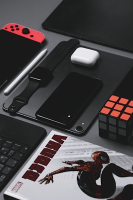 A neatly arranged assortment of tech gadgets and accessories displayed on a gray surface. The layout includes a red and black handheld gaming console, a smartwatch with a black strap, a black smartphone, a white wireless earbud case, a black stylus, and a Rubik's cube. A part of a comic book featuring a superhero is partially visible at the bottom.