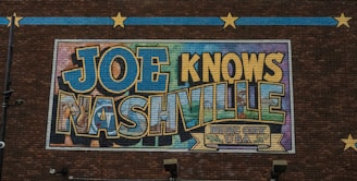 A mural on a brick wall features large, colorful letters spelling out 'JOE KNOWS NASHVILLE' with a decorative banner that reads 'Music City USA' below. The artwork uses a mix of blues, yellows, and greens with an outlined, bold font. Decorative stars and lines on the brick wall add to the visual appeal.
