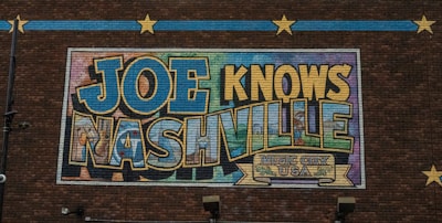 A mural on a brick wall features large, colorful letters spelling out 'JOE KNOWS NASHVILLE' with a decorative banner that reads 'Music City USA' below. The artwork uses a mix of blues, yellows, and greens with an outlined, bold font. Decorative stars and lines on the brick wall add to the visual appeal.