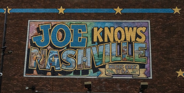 A mural on a brick wall features large, colorful letters spelling out 'JOE KNOWS NASHVILLE' with a decorative banner that reads 'Music City USA' below. The artwork uses a mix of blues, yellows, and greens with an outlined, bold font. Decorative stars and lines on the brick wall add to the visual appeal.