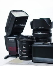A collection of camera equipment including a Canon flash, two camera lenses with visible branding, and a digital camera with an LCD screen. The items are arranged on a flat surface against a plain background.