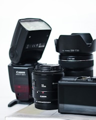 A collection of camera equipment including a Canon flash, two camera lenses with visible branding, and a digital camera with an LCD screen. The items are arranged on a flat surface against a plain background.