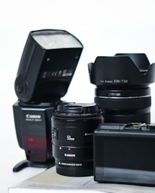 A collection of camera equipment including a Canon flash, two camera lenses with visible branding, and a digital camera with an LCD screen. The items are arranged on a flat surface against a plain background.