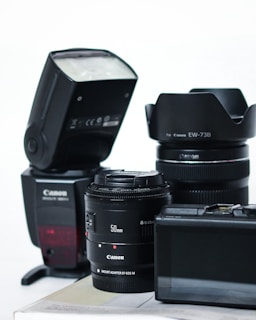 A collection of camera equipment including a Canon flash, two camera lenses with visible branding, and a digital camera with an LCD screen. The items are arranged on a flat surface against a plain background.