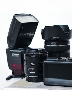A collection of camera equipment including a Canon flash, two camera lenses with visible branding, and a digital camera with an LCD screen. The items are arranged on a flat surface against a plain background.