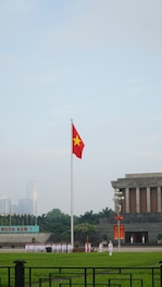 A large flagpole with a red flag featuring a yellow star is prominently displayed in an open area. Below the flag, a group of uniformed individuals stand in formation. To the right, a gray stone building with columns is visible, displaying the name 'Ho Chi Minh' on its facade. The area is surrounded by green lawns and trees, with a distant cityscape in the background.