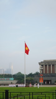 A large flagpole with a red flag featuring a yellow star is prominently displayed in an open area. Below the flag, a group of uniformed individuals stand in formation. To the right, a gray stone building with columns is visible, displaying the name 'Ho Chi Minh' on its facade. The area is surrounded by green lawns and trees, with a distant cityscape in the background.