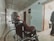 An elderly person in a wheelchair is facing down a long corridor lined with doors. The lighting is dim, casting shadows along the tiled floor. The person is wearing a patterned shirt and the corridor appears to be in a clinical or institutional setting, possibly a hospital or care facility. An open door on the right side reveals part of another room.