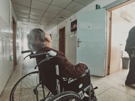 An elderly person in a wheelchair is facing down a long corridor lined with doors. The lighting is dim, casting shadows along the tiled floor. The person is wearing a patterned shirt and the corridor appears to be in a clinical or institutional setting, possibly a hospital or care facility. An open door on the right side reveals part of another room.