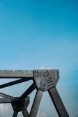 A close-up view of a metallic structural component with rivets against a clear blue sky. The structure has angular lines and industrial design, suggesting part of a bridge or architectural framework.