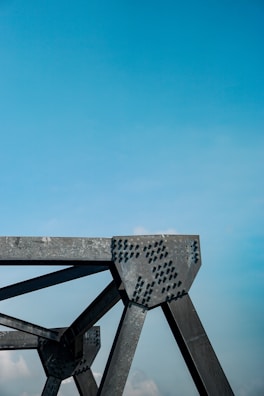 A close-up view of a metallic structural component with rivets against a clear blue sky. The structure has angular lines and industrial design, suggesting part of a bridge or architectural framework.