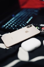 A metallic iPhone with a dual camera is placed next to a white wireless earbud case, atop a black and white fabric surface. In the background, a laptop keyboard with illuminated keys is partially visible.