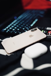 A metallic iPhone with a dual camera is placed next to a white wireless earbud case, atop a black and white fabric surface. In the background, a laptop keyboard with illuminated keys is partially visible.