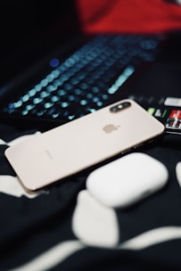A metallic iPhone with a dual camera is placed next to a white wireless earbud case, atop a black and white fabric surface. In the background, a laptop keyboard with illuminated keys is partially visible.