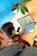 A person is using a laptop on a wooden desk, viewing a map displayed on the screen. Another person is sitting nearby, partially visible. A smartphone and a pot with a green plant are also on the desk. The background is a bright blue wall.