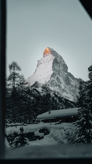 A majestic snow-capped mountain peak with the sun illuminating the tip, surrounded by dark, dense pine trees. A cozy cabin sits nestled in the snowy landscape.