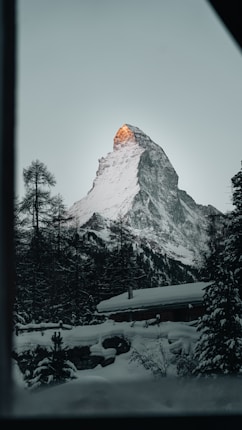 A majestic snow-capped mountain peak with the sun illuminating the tip, surrounded by dark, dense pine trees. A cozy cabin sits nestled in the snowy landscape.
