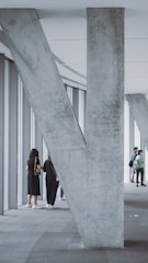 A modern architectural space featuring a large, V-shaped concrete pillar supporting the ceiling. Several people stand near the windows, some looking out while others converse. The floor is tiled, and the space has an open, airy feel with natural light streaming in from the windows.
