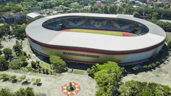An aerial view of a large oval stadium with a grey roof and colorful outer walls. The stadium is surrounded by lush green trees and a few buildings. There is a circular area with a statue and pathways leading to the stadium.
