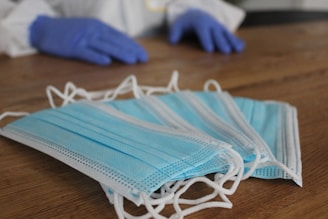 Several blue disposable face masks are laid out on a wooden surface. A pair of hands wearing blue gloves can be seen in the background.