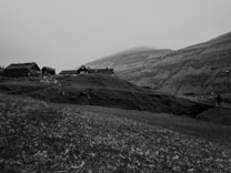 A rural landscape features a group of rustic buildings, likely farmhouses, situated on a grassy hillside. The terrain is steep, with a backdrop of undulating hills partially obscured by fog. The area appears remote and tranquil, with no visible human presence.