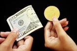 Two hands are visible against a black background. One hand holds a $50 US bill, while the other holds a gold-colored Bitcoin coin.