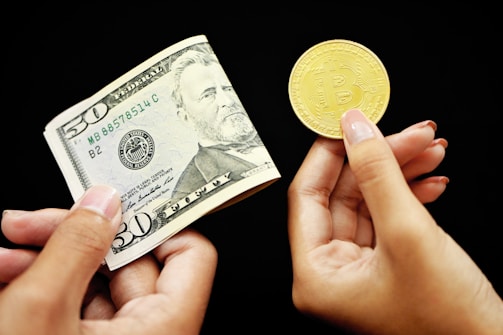 Two hands are visible against a black background. One hand holds a $50 US bill, while the other holds a gold-colored Bitcoin coin.