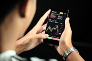 A person is holding a smartphone displaying a financial trading app. The screen shows a candlestick chart with various trading metrics, including the current price, percentage changes, and buttons for different actions. The person's hands are visible, and a black background accentuates the smartphone screen.