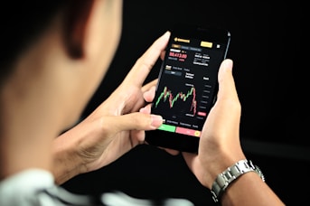 A person is holding a smartphone displaying a financial trading app. The screen shows a candlestick chart with various trading metrics, including the current price, percentage changes, and buttons for different actions. The person's hands are visible, and a black background accentuates the smartphone screen.