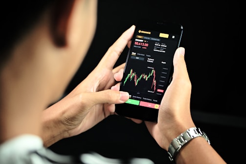 A person is holding a smartphone displaying a financial trading app. The screen shows a candlestick chart with various trading metrics, including the current price, percentage changes, and buttons for different actions. The person's hands are visible, and a black background accentuates the smartphone screen.