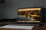 A laptop displaying a scenic desktop wallpaper with rugged coastlines and mountains viewed from a side angle. The scene is set on a wooden desk with a keyboard in the foreground. The overall setting gives off a minimalist and modern vibe.