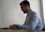 A person is seated at a wooden table, working on a laptop. They appear to be smiling, suggesting enjoyment or satisfaction. The background is a plain, light-colored wall, providing a simple and neutral setting.