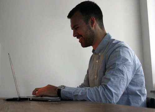 A person is seated at a wooden table, working on a laptop. They appear to be smiling, suggesting enjoyment or satisfaction. The background is a plain, light-colored wall, providing a simple and neutral setting.