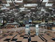 A spacious and modern airport terminal with a high, geometric-patterned ceiling and a shiny tiled floor. In the center, there are flight information displays, surrounded by automated check-in kiosks and counters. The background features a green wall with plants, adding to the refreshing atmosphere. The area is well-lit and appears organized.