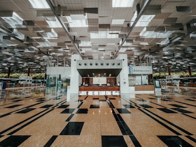 A spacious and modern airport terminal with a high ceiling featuring geometric designs and a clean, polished floor with black and tan tiles. The center has an information counter with signs and electronic displays, and various airport amenities are visible in the background.