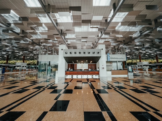 A spacious and modern airport terminal with a high ceiling featuring geometric designs and a clean, polished floor with black and tan tiles. The center has an information counter with signs and electronic displays, and various airport amenities are visible in the background.