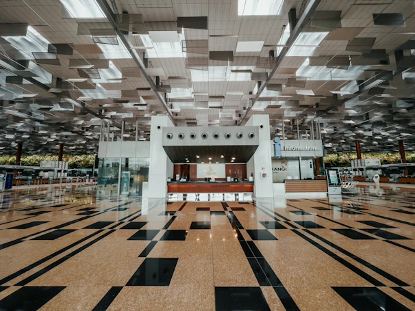 A spacious and modern airport terminal with a high ceiling featuring geometric designs and a clean, polished floor with black and tan tiles. The center has an information counter with signs and electronic displays, and various airport amenities are visible in the background.