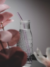 A clear glass bottle with a purple straw stands on a reflective surface with droplets of water on its exterior. The design is minimalistic and elegant, surrounded by blurred leaves in soft pink tones, creating a peaceful ambiance.