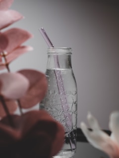 A clear glass bottle with a purple straw stands on a reflective surface with droplets of water on its exterior. The design is minimalistic and elegant, surrounded by blurred leaves in soft pink tones, creating a peaceful ambiance.
