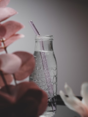 A clear glass bottle with a purple straw stands on a reflective surface with droplets of water on its exterior. The design is minimalistic and elegant, surrounded by blurred leaves in soft pink tones, creating a peaceful ambiance.