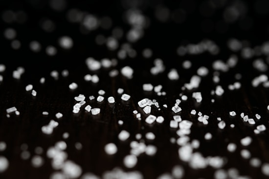 Small white crystals scattered over a dark surface, with a soft focus and some areas highlighted. The crystals are various shapes and sizes, creating a textured appearance against the black background.