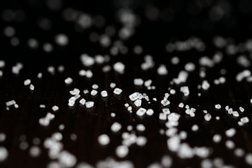 Small white crystals scattered over a dark surface, with a soft focus and some areas highlighted. The crystals are various shapes and sizes, creating a textured appearance against the black background.