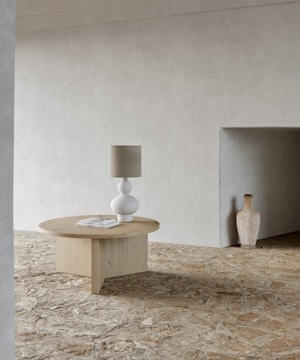 A minimalist interior features a round wooden table with a white, spherical-based lamp on top. A few stacked books lie near the lamp. The space has a textured, light-colored wall and a rustic stone floor. In the background, a ceramic vase is placed next to an inset in the wall.
