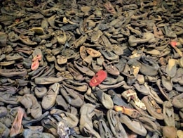 A large pile of old, worn-out shoes with various styles and sizes scattered haphazardly. Some shoes have visible wear and tear, with frayed edges and scuffed surfaces. The shoes vary slightly in color, with a generally dark and muted palette.